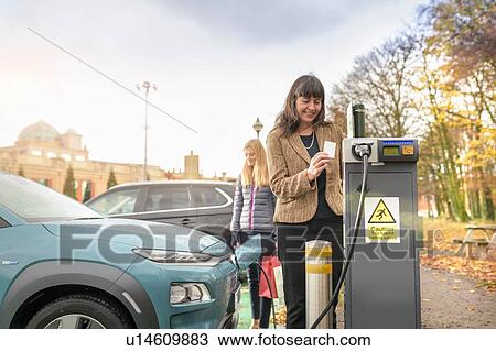 Stock Image - Female shoppers charging electric car at charge point, Manchester, UK. Fotosearch