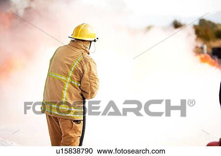 Stock Image - Firemen training, fireman spraying firefighting foam at training facility. Fotosearch