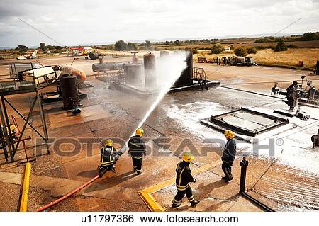 Stock Photograph - Firemen training, firemen spraying firefighting foam at oil storage tank at training facility. Fotosearch