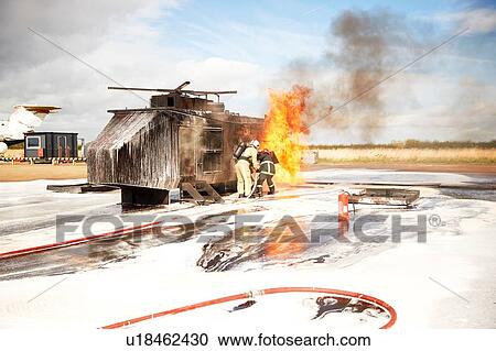 Stock Image - Firemen training, team of firemen spraying firefighting foam at mock helicopter fire at training facility. Fotosearch
