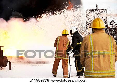 Stock Photo - Firemen training, team of firemen spraying firefighting foam on fire at training facility, rear view. Fotosearch