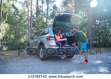 Stock Image - Male and female runners tying laces on car boot in sunlit forest. Fotosearch
