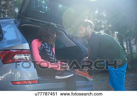 Stock Image - Male and female runners tying laces on car boot in sunlit forest. Fotosearch