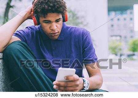Stock Photo - Man texting and using headphones on concrete bench. Fotosearch