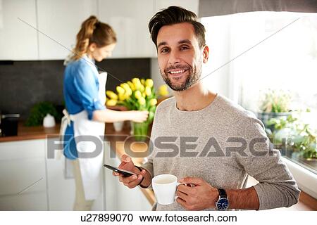 Man using smartphone in kitchen, woman arranging flowers in background View Large Photo Image Stock Image - Man using smartphone in kitchen, woman arranging flowers in background. Fotosearch