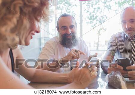 Picture - Mid adult woman and male friends looking at smartphones in cafe. Fotosearch