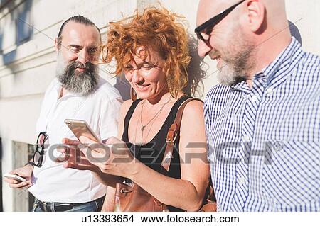 Mid adult woman and male friends leaning against wall looking at smartphone View Large Photo Image Picture - Mid adult woman and male friends leaning against wall looking at smartphone. Fotosearch