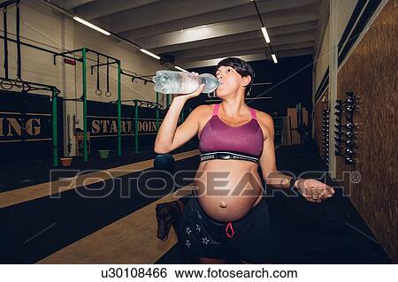 Pregnant woman drinking from water bottle in gym View Large Photo Image Stock Photograph - Pregnant woman drinking from water bottle in gym. Fotosearch