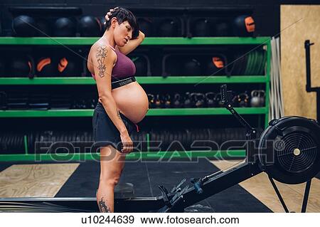 Stock Photo - Pregnant woman looking at rowing machine in gym. Fotosearch
