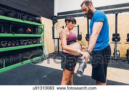 Stock Image - Pregnant woman using bar bell in gym. Fotosearch
