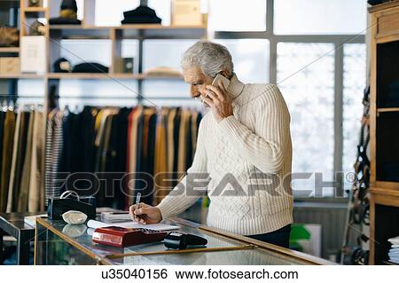 Stock Photograph - Senior man working in clothes shop using mobile phone. Fotosearch