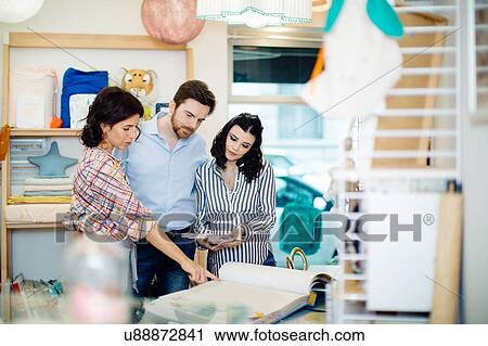 Stock Image - Shop keeper showing pregnant couple sample book. Fotosearch