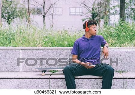 Stock Image - Skateboarder using headphones on concrete bench, Milan, Italy. Fotosearch