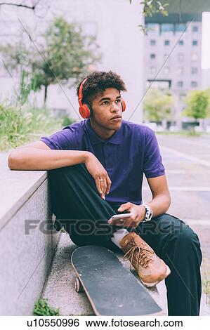 Stock Photograph - Skateboarder using headphones on concrete bench, Milan, Italy. Fotosearch