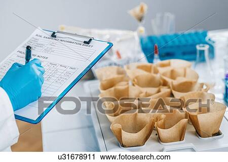 Stock Image - Soil testing laboratory. Close up image of female scientist wearing white coat taking notes in laboratory. Fotosearch