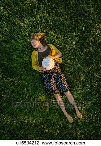 Stock Image - Teenage girl lying on grass with eyes closed holding soccer ball, overhead view. Fotosearch