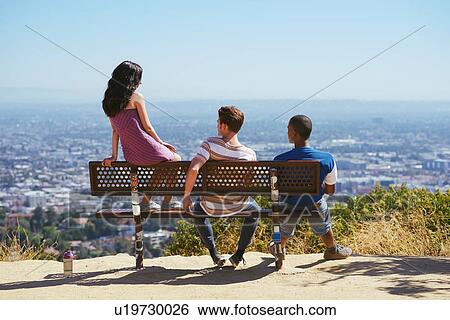 Stock Photograph - Three young adult friends looking out at cityscape from hilltop bench, rear view, Los Angeles, California, USA. Fotosearch