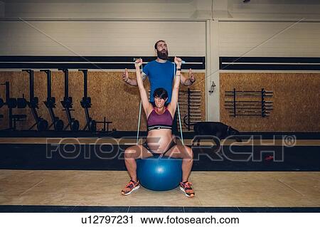 Stock Image - Trainer guiding pregnant woman using ropes in gym. Fotosearch
