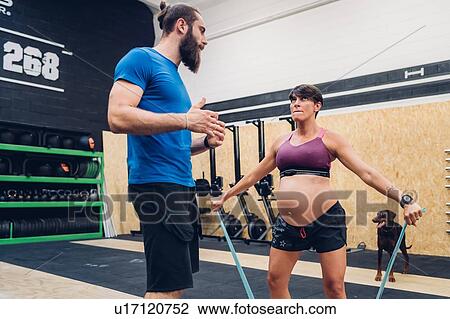 Stock Image - Trainer guiding pregnant woman using ropes in gym. Fotosearch