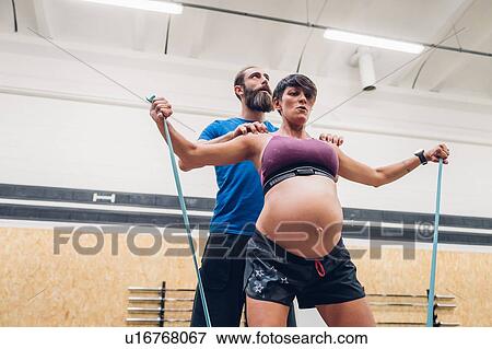 Stock Photo - Trainer guiding pregnant woman using ropes in gym. Fotosearch