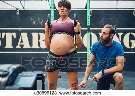 Stock Photo - Trainer guiding pregnant woman using weights in gym. Fotosearch