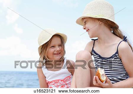 Stock Image - Two girls in sun hats eating sandwiches on beach, Scopello, Sicily, Italy. Fotosearch
