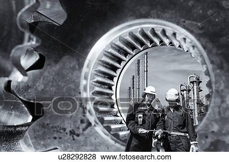 Stock Photo - Two workers and giant gear axle in foreground with an industrial plant in the background.. Fotosearch