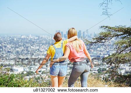 Stock Image - Two young female friends looking at skyline from hilltop, rear view, Los Angeles, California, USA. Fotosearch