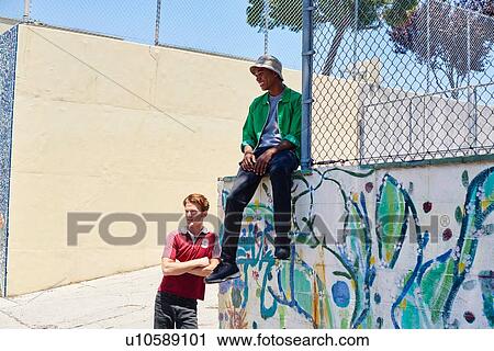 Stock Image - Two young men waiting by park fence, Los Angeles, California, USA. Fotosearch