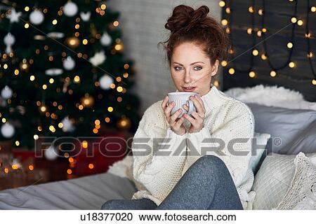 Stock Image - Woman having warm drink on bed decorated with Christmas lights. Fotosearch