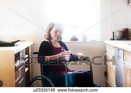 Stock Photograph - Woman in wheelchair with glass of water in kitchen. Fotosearch