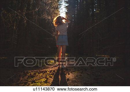 Stock Image - Woman looking up from dark sunlit forest, rear view. Fotosearch