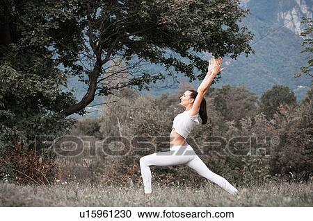 Stock Image - Woman practicing yoga, warrior pose in rural landscape. Fotosearch