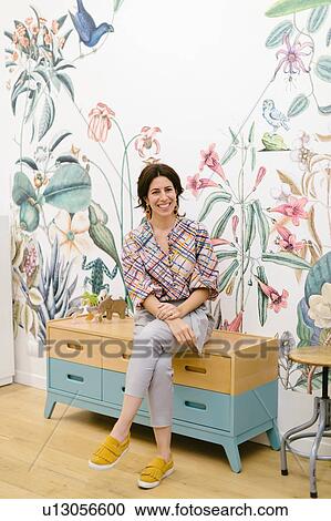 Stock Image - Woman sitting on cabinet, floral pattern wallpaper in background. Fotosearch