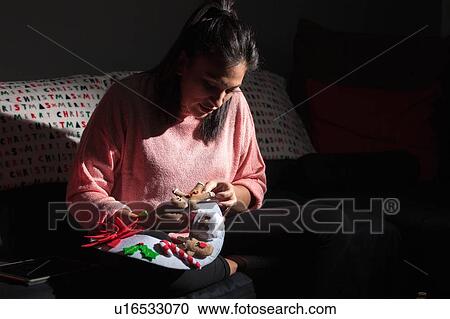 Stock Image - Woman sitting on sofa stitching felt gingerbread man onto christmas decoration. Fotosearch
