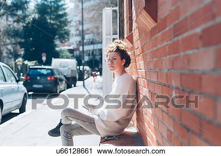 Stock Image - Woman with cellphone on bench on sunny street, Milan, Italy. Fotosearch