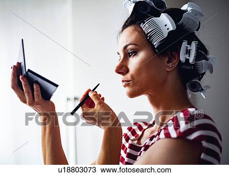 Stock Image - Woman with hair-rollers putting on make up. Fotosearch