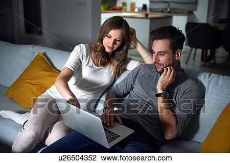 Stock Image - Young couple sitting on sofa in evening looking at laptop. Fotosearch