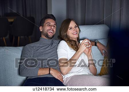 Young couple sitting on sofa in evening, laughing while watching TV View Large Photo Image Picture - Young couple sitting on sofa in evening, laughing while watching TV. Fotosearch