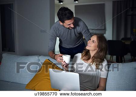 Young man handing coffee to girlfriend sitting on sofa in evening View Large Photo Image Stock Image - Young man handing coffee to girlfriend sitting on sofa in evening. Fotosearch
