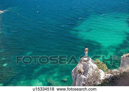Stock Photo - Young man looking out from coastal rock, Cagliari, Sardinia, Italy. Fotosearch