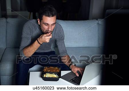 Stock Image - Young man sitting on sofa in evening eating takeaway and using laptop. Fotosearch