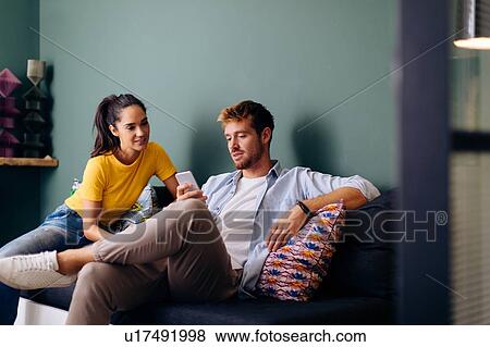 Stock Photo - Young woman looking at man using smartphone on couch. Fotosearch