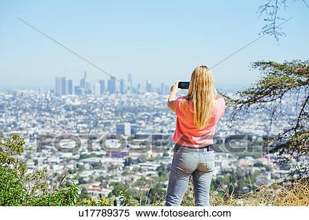 Stock Photography - Young woman photographing skyline from hilltop, rear view, Los Angeles, California, USA. Fotosearch