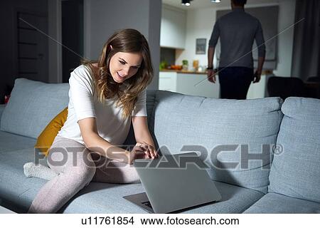 Young woman sitting on sofa in evening, peeking at boyfriend's laptop View Large Photo Image Picture - Young woman sitting on sofa in evening, peeking at boyfriend's laptop. Fotosearch