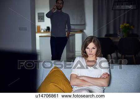 Young woman sulking on sofa in evening, while boyfriend makes smartphone call View Large Photo Image Stock Image - Young woman sulking on sofa in evening, while boyfriend makes smartphone call. Fotosearch