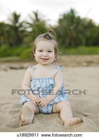 Stock Image - Baby girl wearing blue dress sitting on beach. Fotosearch