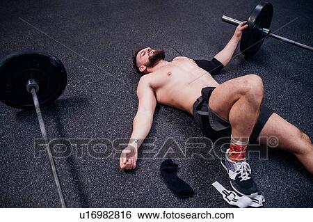 Stock Photograph - Bare chested young man training, lying exhausted between barbells in gym. Fotosearch
