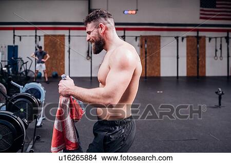 Picture - Bare chested young man training, holding bottled water and towel in gym, side view. Fotosearch