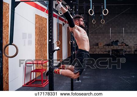 Stock Image - Bare chested young man training, swinging on exercise bar in gym. Fotosearch